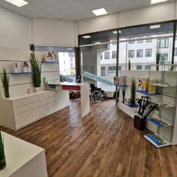 Innenansicht eines Geschäfts mit weißen Regalen, Pflanzen und einem Rollstuhl im Hintergrund.