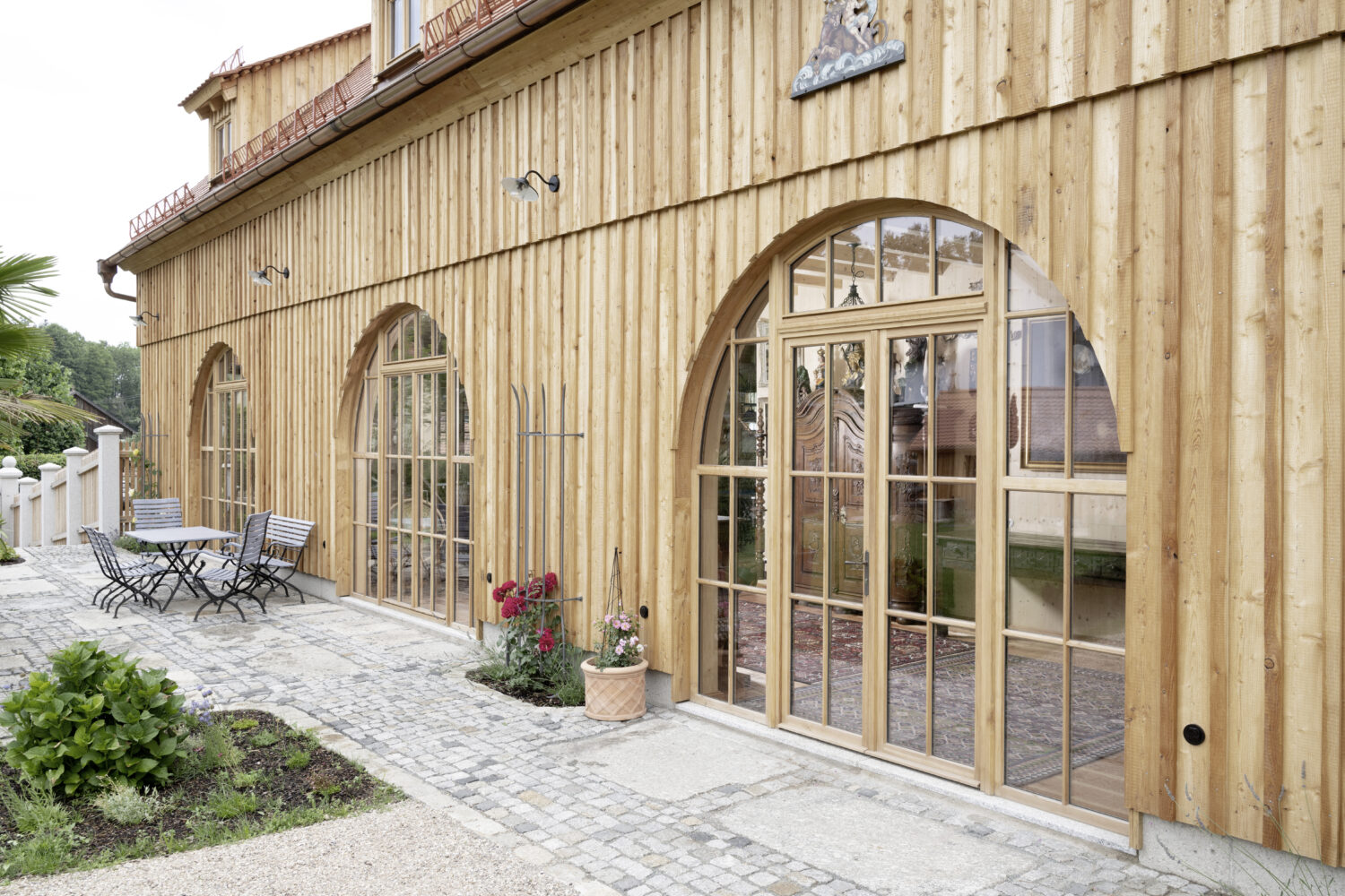 Außenansicht eines Gebäudes mit vertikaler Holzfassade und großen, maßgefertigten Holzfenstern und Türen mit Rundbogen. Vor der Fassade befindet sich eine gepflasterte Terrasse mit Sitzbereich.