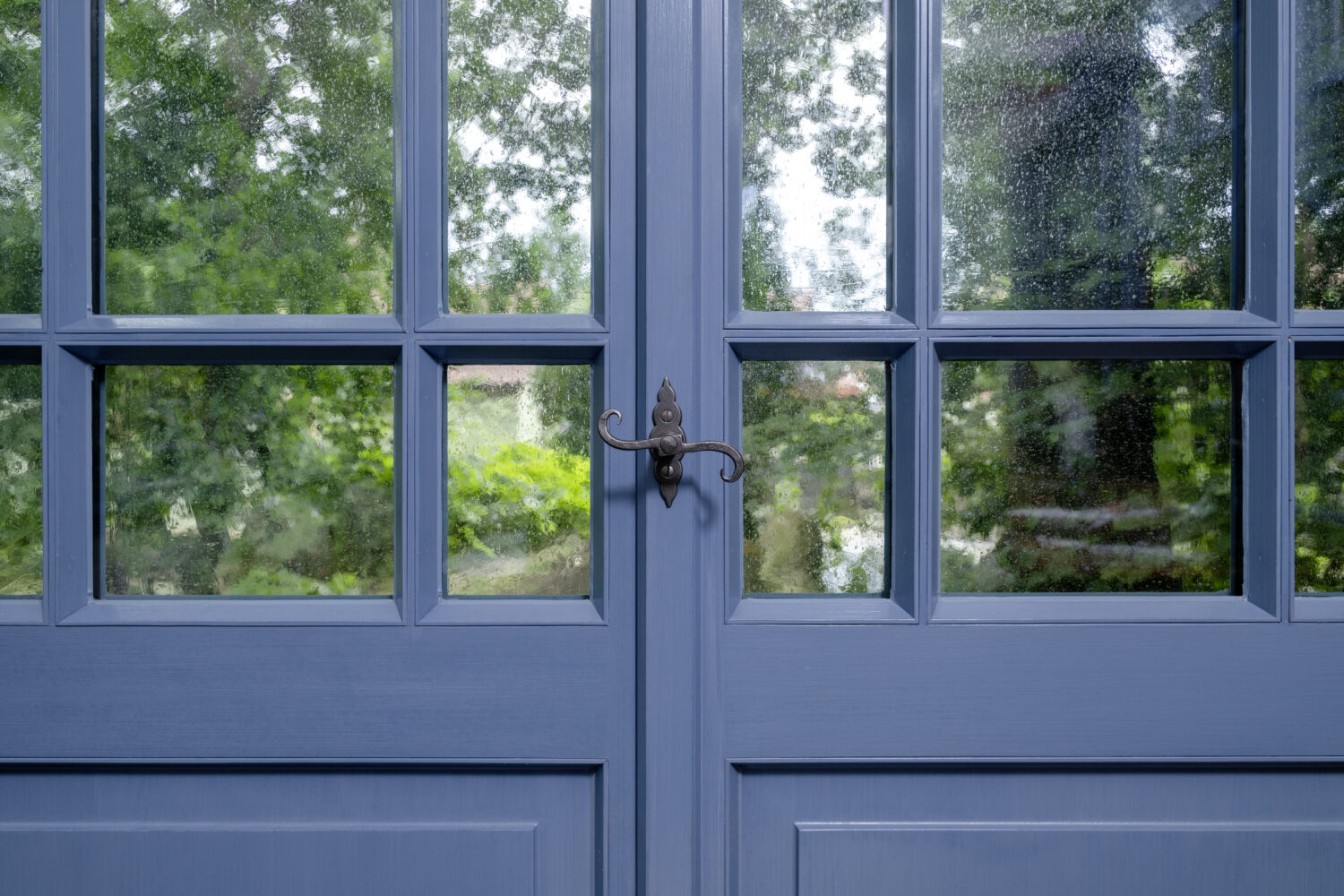 Detailaufnahme einer blauen Holzfensterfront mit Sprossen und schmiedeeisernem Griff. Durch die strukturierten Glasscheiben sind unscharfe grüne Bäume im Hintergrund sichtbar